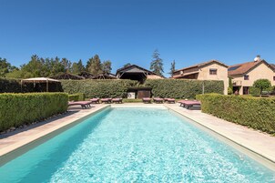 A rectangular swimming pool with lounge chairs and a covered area on one side, surrounded by hedges and trees.