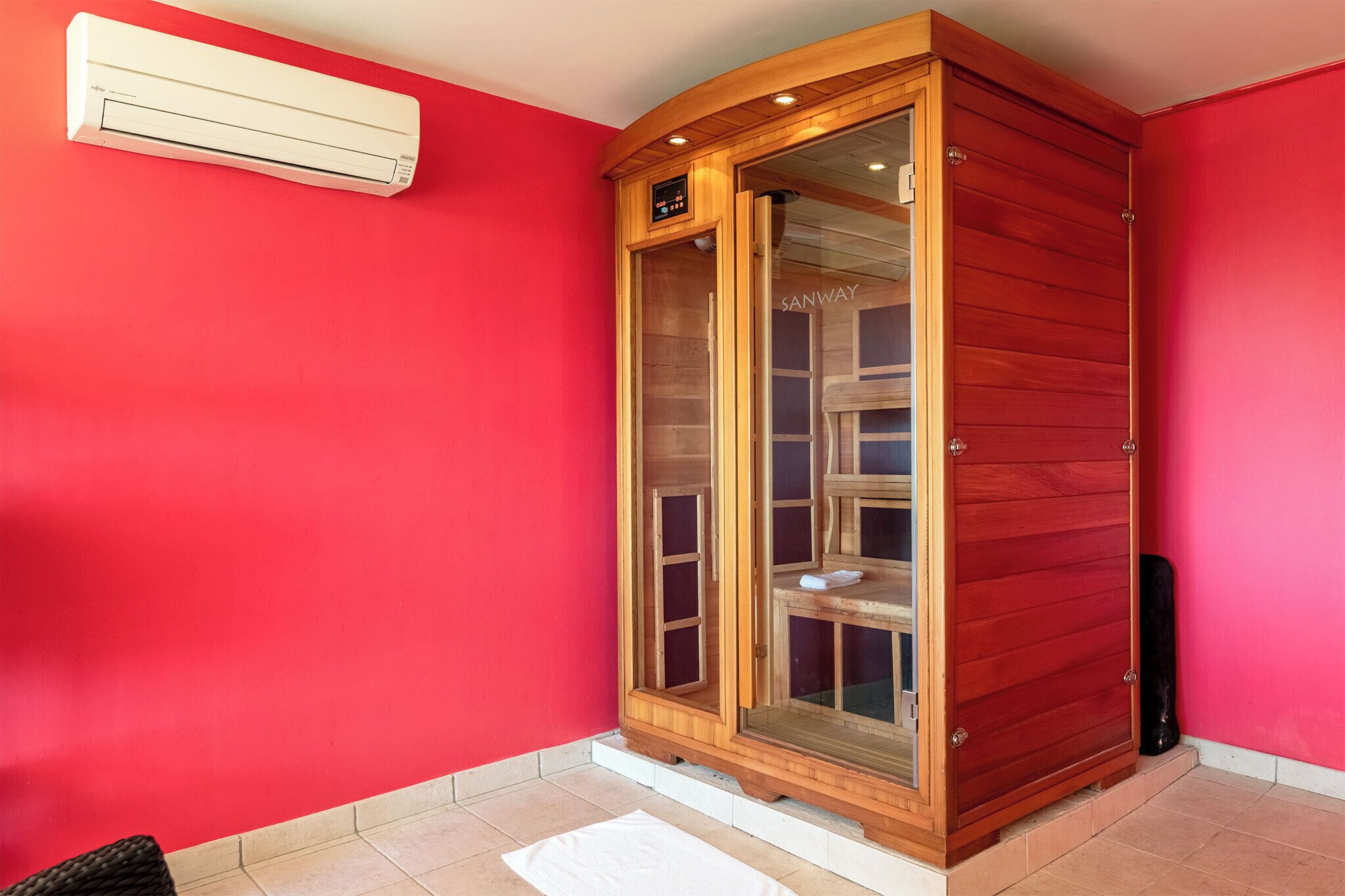 Een sauna met een houten buitenkant en glazen deuren, rode wanden en een witte airconditioning unit erboven.