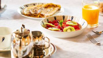 Une table mise pour le petit-déjeuner avec un saladier de fruits frais, un verre de jus d’orange et une assiette de crêpes.