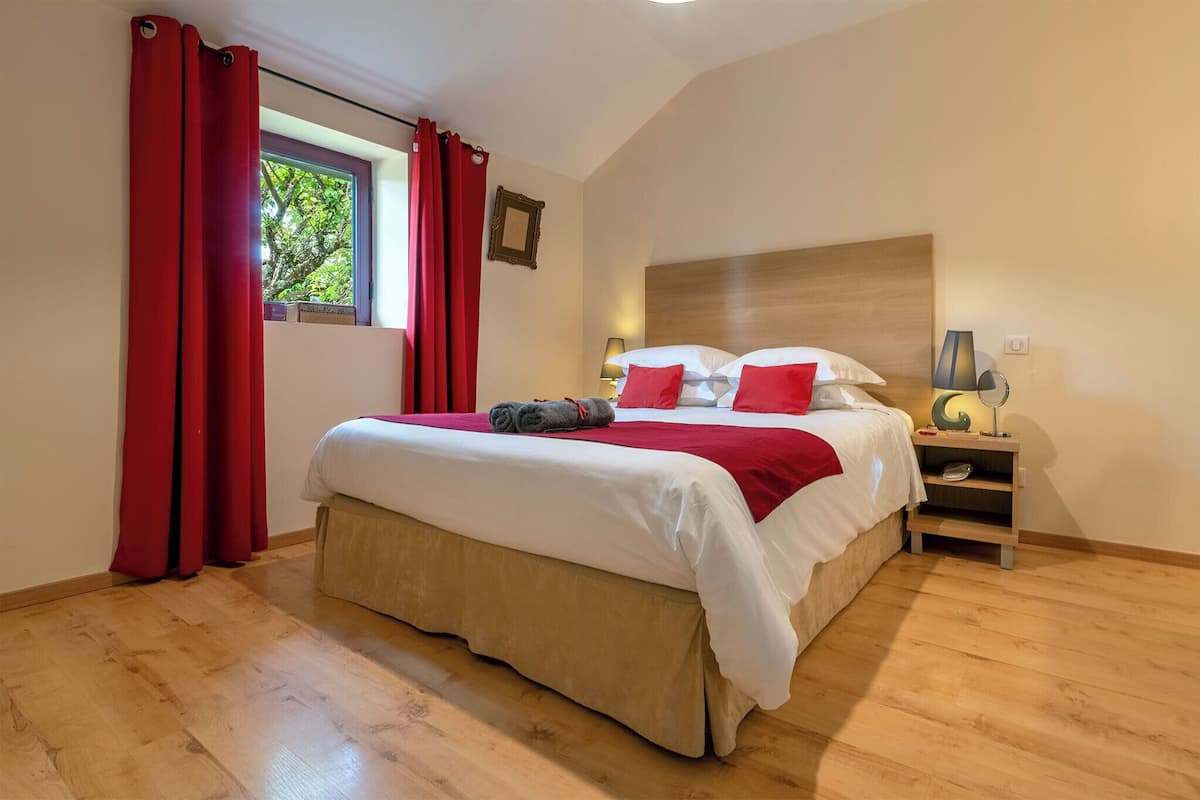 Un dormitorio con una cama grande, cortinas rojas, una mesita de noche con lámpara y una ventana con vistas a árboles.