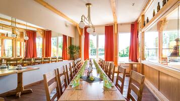 A long dining table with chairs, wooden beams, and large windows with red curtains.