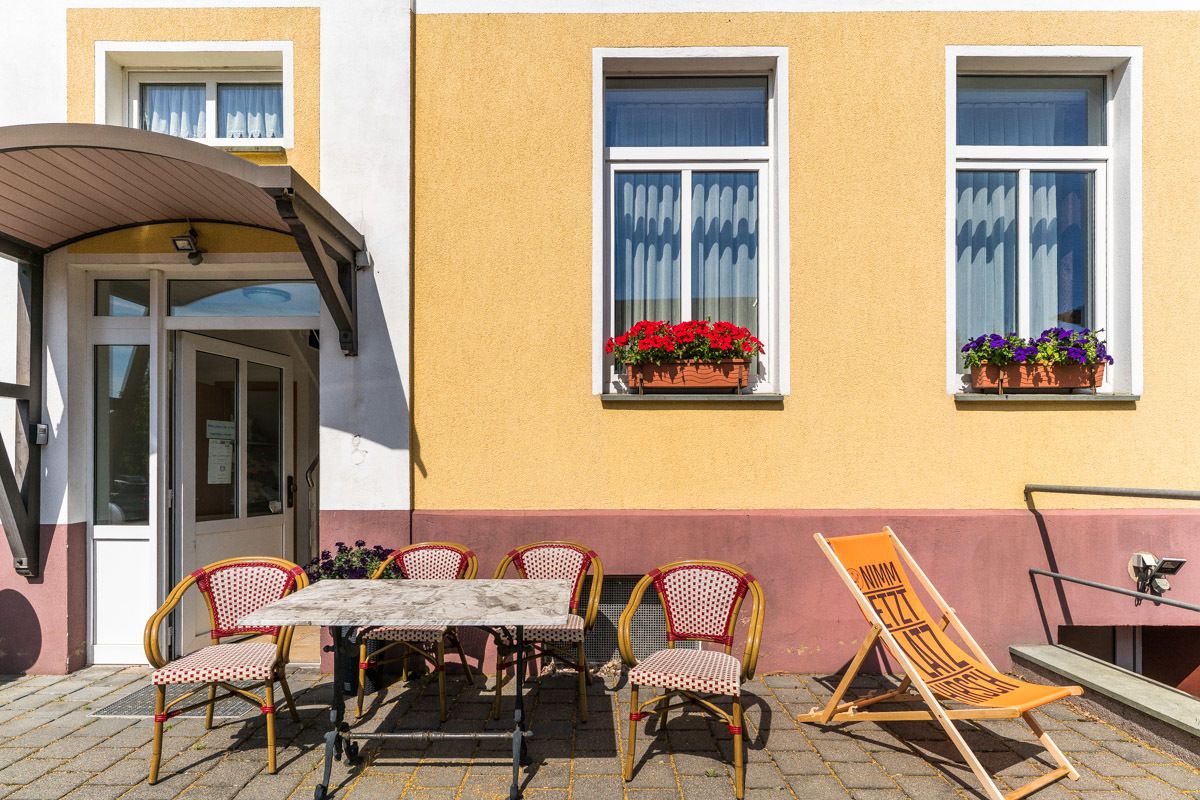 Ein Gebäude mit gelber Fassade, einer überdachten Eingangspartie, einem Tisch mit Stühlen und Blumentöpfen an den Fensterbänken.