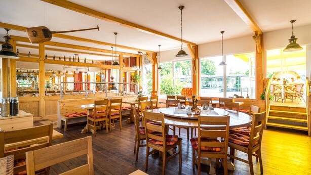 Un interior rústico de madera con mesas y sillas redondas, una zona de bar con estanterías y amplios ventanales que dejan entrar luz natural.