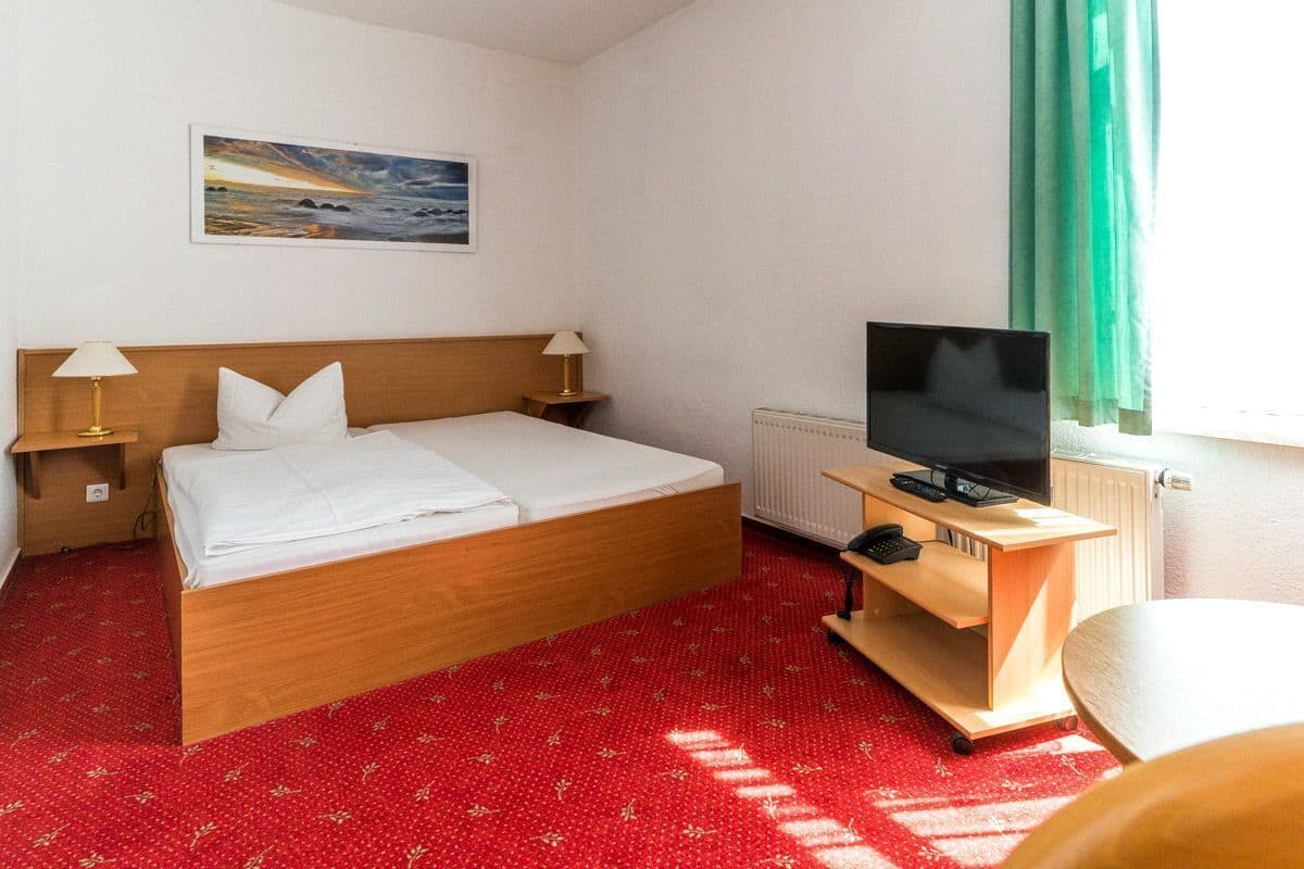 Une chambre d’hôtel avec un lit, deux lampes de chevet, un téléviseur posé sur un meuble, une fenêtre avec des rideaux et un tableau accroché au mur.