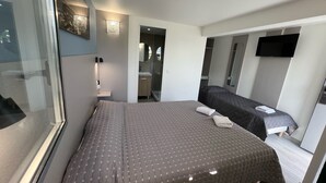 Quarto de hotel moderno com uma cama grande, mesa de cabeceira, espelho, TV fixada na parede e banheiro visível através de uma porta aberta.