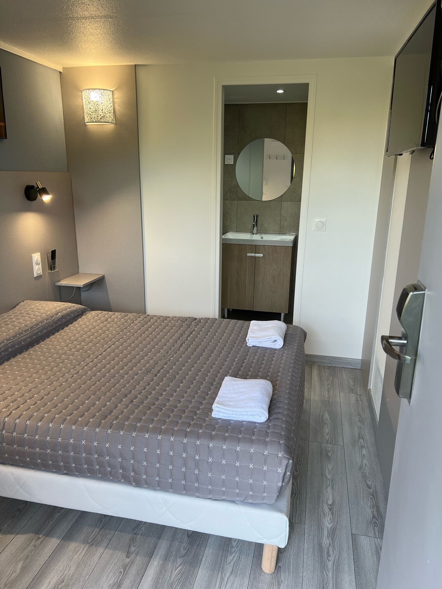 A bedroom with a large bed, a bedside table, a wall-mounted light, a mirror, and a bathroom with a sink and mirror.