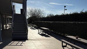 Área externa iluminada pelo sol, com escadaria, mesas de piquenique e um viveiro.