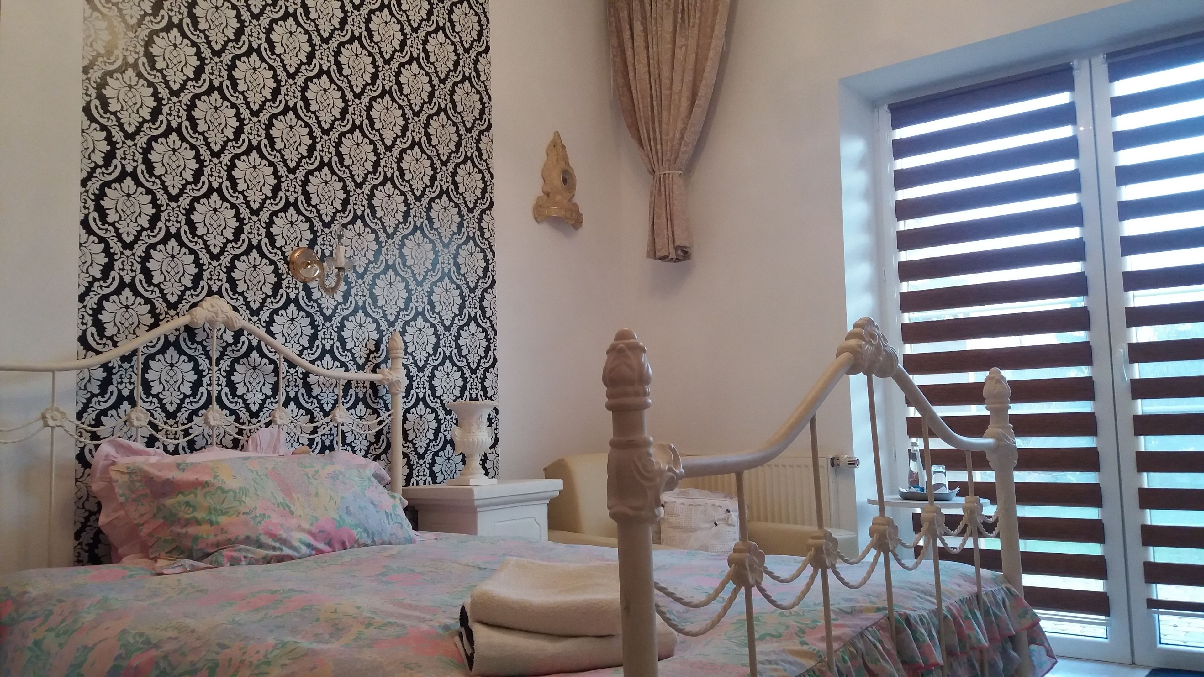 A bedroom with a four-poster bed, a patterned wallpaper, a window with blinds, and a nightstand.