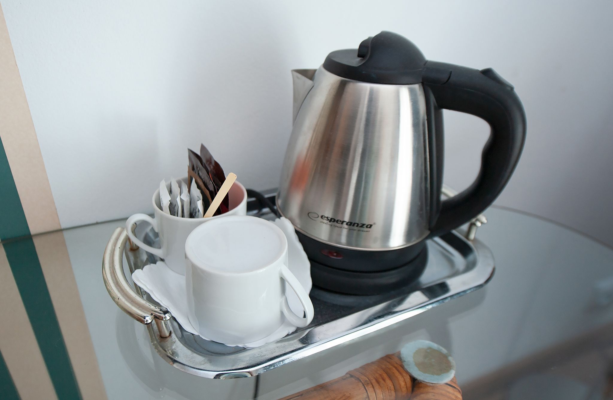 Una tetera eléctrica de acero inoxidable con asa negra y una taza blanca con una bolsita de té dentro, sobre una bandeja reflectante.