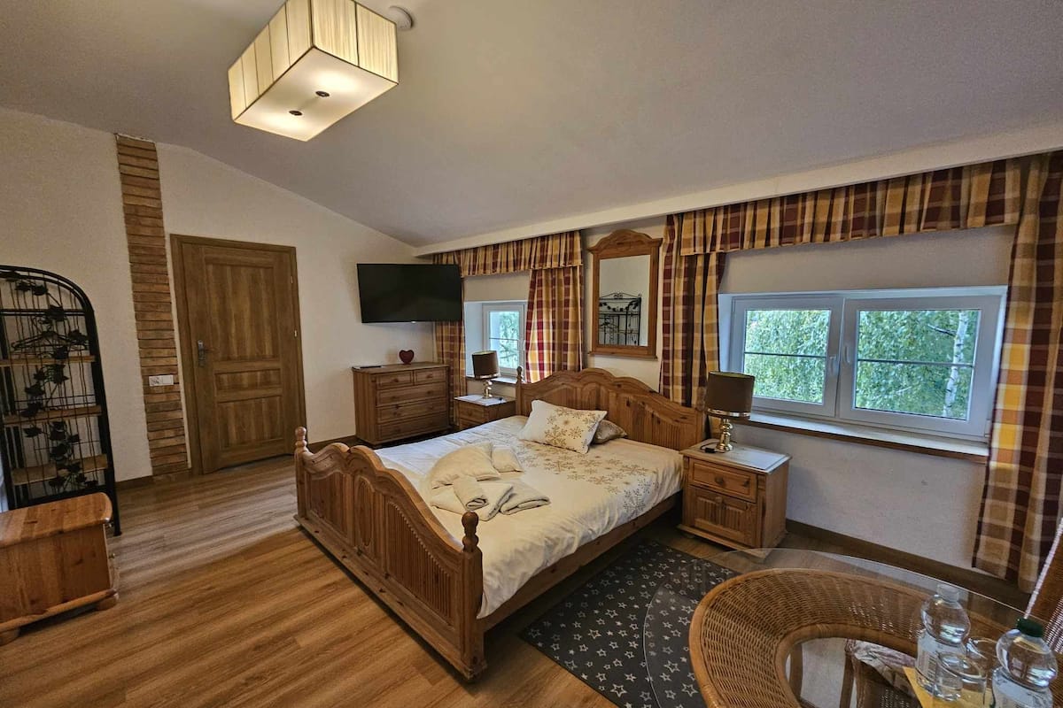 A bedroom with a wooden bed, a television, a window with curtains, a bedside table, and a chair.