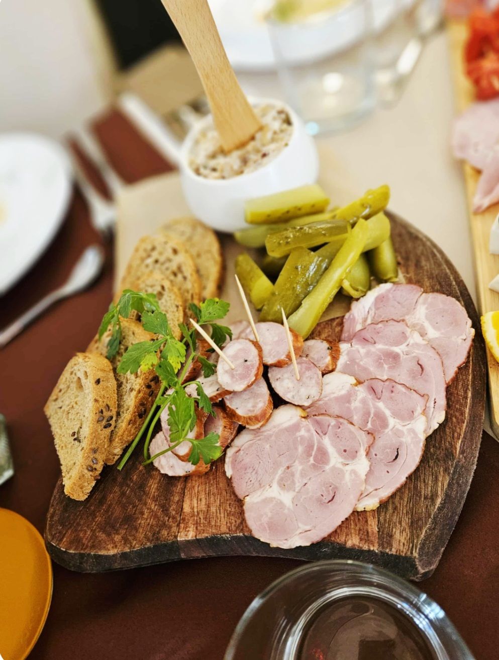 Una tabla de madera con jamón en rodajas, pepinillos y pan sobre una mesa.