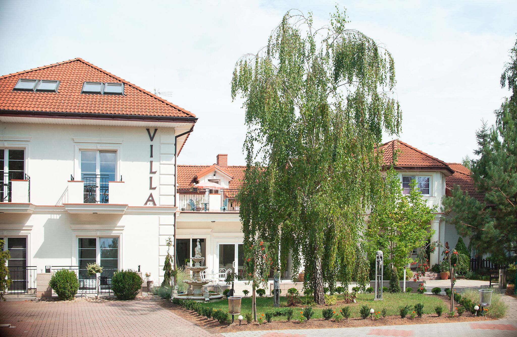 Eine Villa mit Ziegeldach, dem Namen 'VILLA', der sichtbar ist, und einem gepflegten Garten mit Brunnen und verschiedenen Pflanzen.