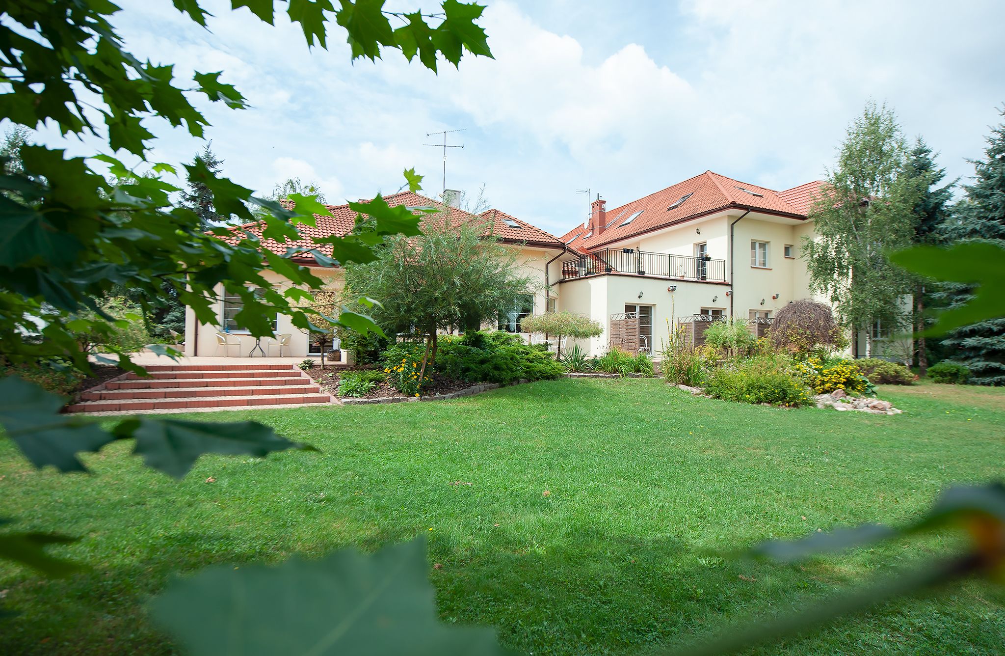 A two-story house with a red-tiled roof, a front garden, and a staircase leading to the entrance.