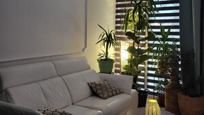 Una sala moderna con un sofá blanco, una mesa de centro redonda y varias plantas en macetas. Un letrero en la pared dice 'Villa Hubertus Boutique Design'.