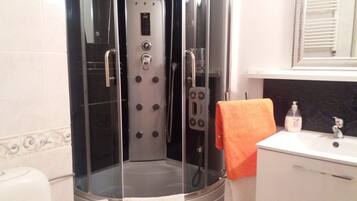 A glass-enclosed shower stall with a control panel, a white sink with a soap dispenser, and a towel rack with an orange towel.