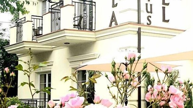 Una villa de dos pisos con un balcón, un cartel que dice "VILLA HUBERTUS HOTEL" y rosas rosadas en primer plano.