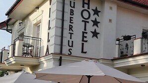 Un hotel llamado "HUBERTUS" de estilo arquitectónico clásico, con un balcón que tiene barandilla y plantas en macetas.