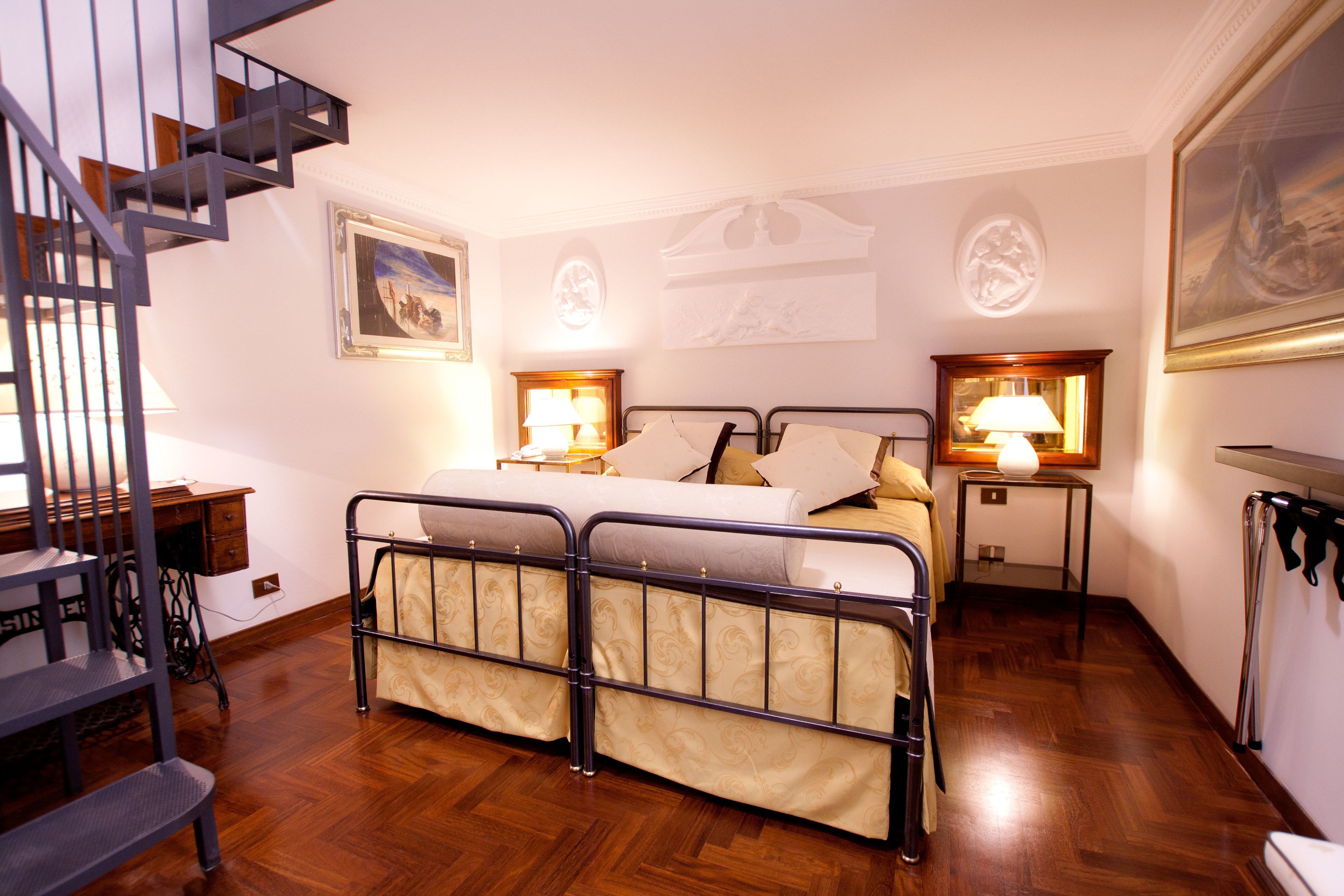 A bedroom with a double bed, two bedside tables with lamps, a wooden desk, a chair, a staircase, and a painting on the wall.