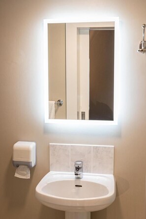 A bathroom with a white sink, a mirror with a light, and a paper towel dispenser. - Tralee Townhouse (Tralee)