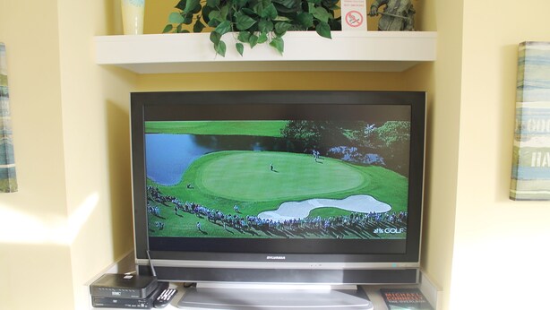 Een televisie waarop een golfbaan te zien is, een afstandsbediening en een boek met de titel 'The Overlook' op een plank.