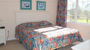Een slaapkamer met een bloemenbedekker, een witte dressoir, een nachtkastje en een raam met uitzicht op groen.
