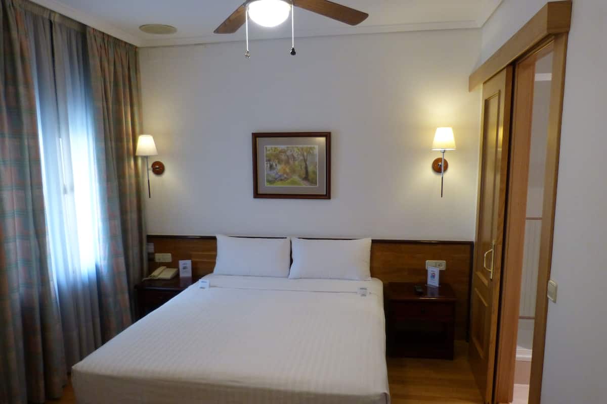 Une chambre d’hôtel avec un grand lit, deux tables de chevet, un ventilateur de plafond, un tableau au mur et une porte sur le côté droit.