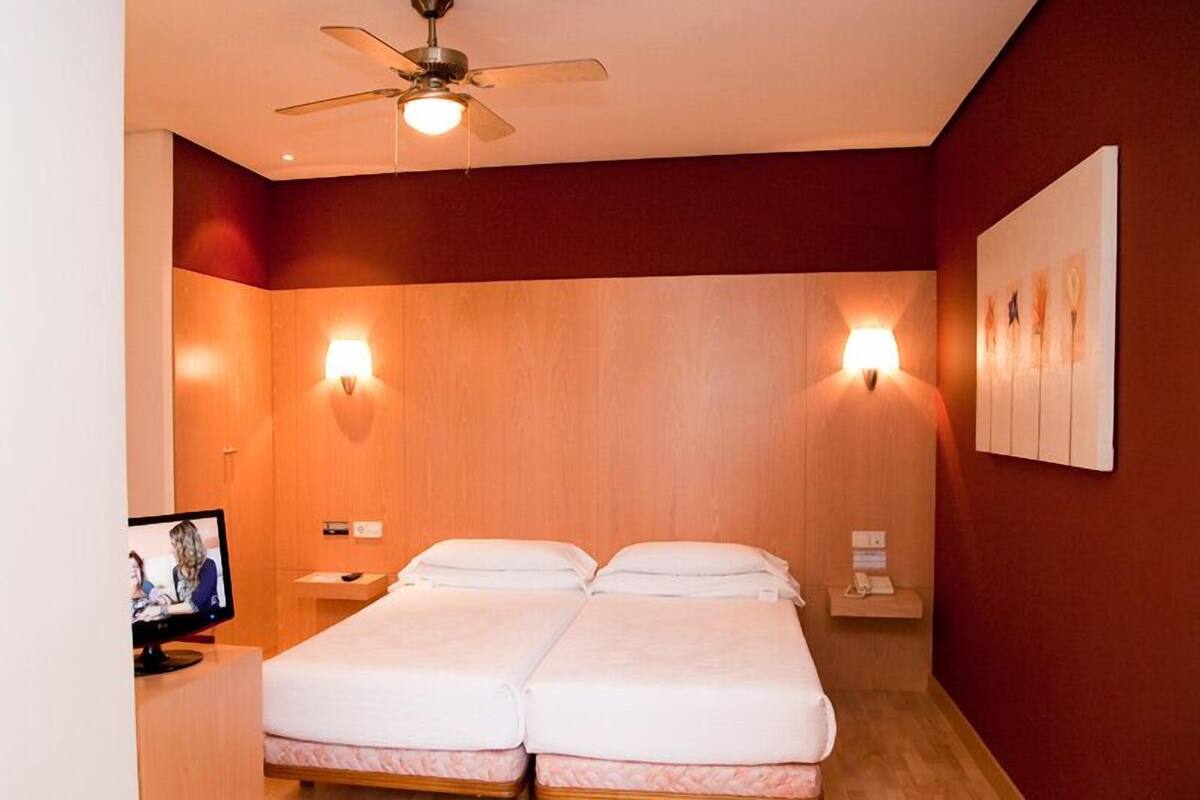 Une chambre d’hôtel avec un lit double, deux tables de chevet, un ventilateur de plafond et deux lampes fixées au mur.