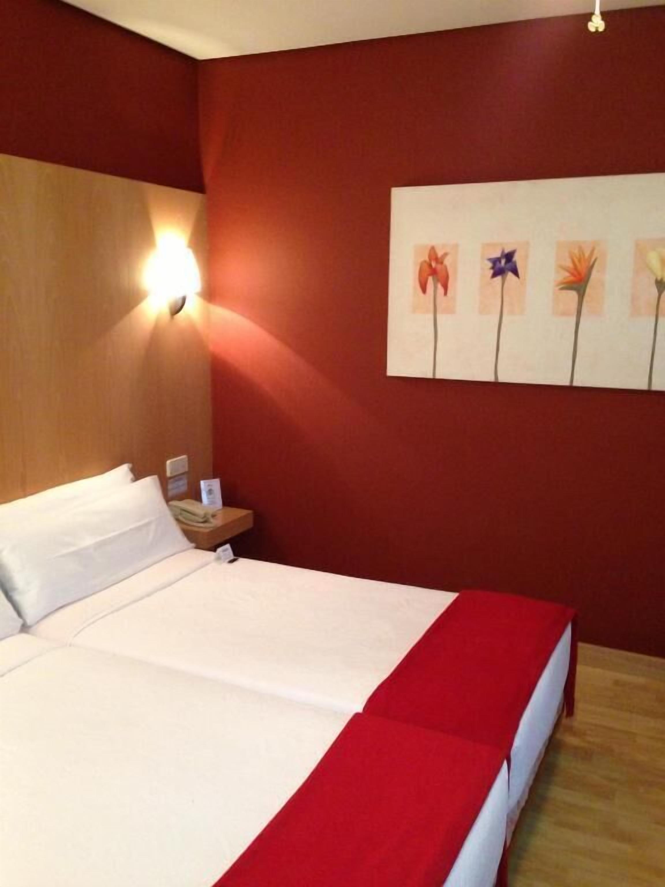 A hotel room with a bed, a nightstand, a wall-mounted light, and a floral artwork.
