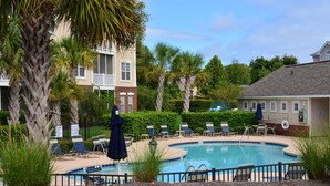 A pool area with lounge chairs and umbrellas, surrounded by palm trees and a building with a tiled roof. - 123 Harbour Cove (North Myrtle Beach)