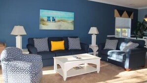 A living room with a blue wall, two sofas with patterned cushions, a white coffee table, and a painting of a beach scene. - 1515 The Havens (North Myrtle Beach)