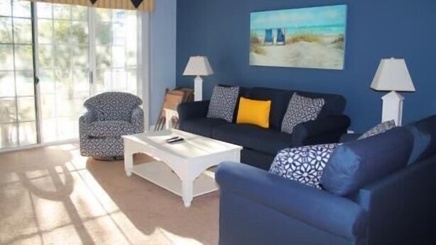 A living room with a blue sofa, patterned armchair, and a coffee table. There are two lamps and a painting of a beach scene on the wall. - 1515 The Havens (North Myrtle Beach)
