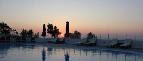 A swimming pool with lounge chairs and umbrellas, set against a sunset sky.