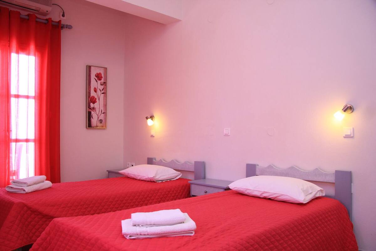 Habitación de hotel con dos camas, ropa de cama roja, una cortina roja, un aire acondicionado instalado en la pared y un cuadro enmarcado.