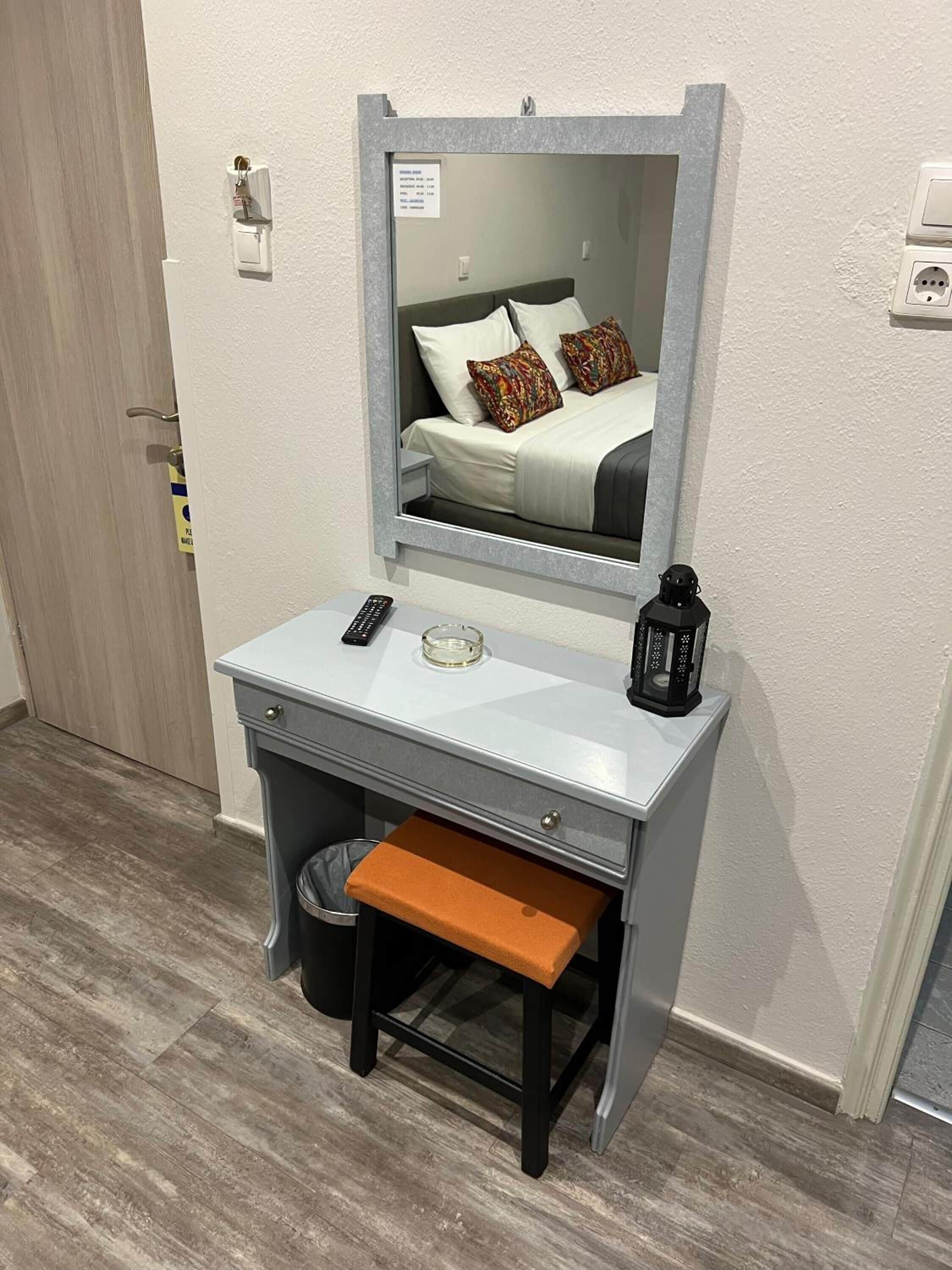 Une chambre d’hôtel compacte comprenant un bureau gris, un petit tabouret, un miroir, une télécommande, un verre et une lanterne.