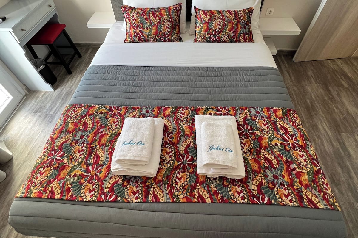 Una cama doble con colchón gris, dos almohadas blancas y una colcha floral de colores. Hay dos toallas con el nombre 