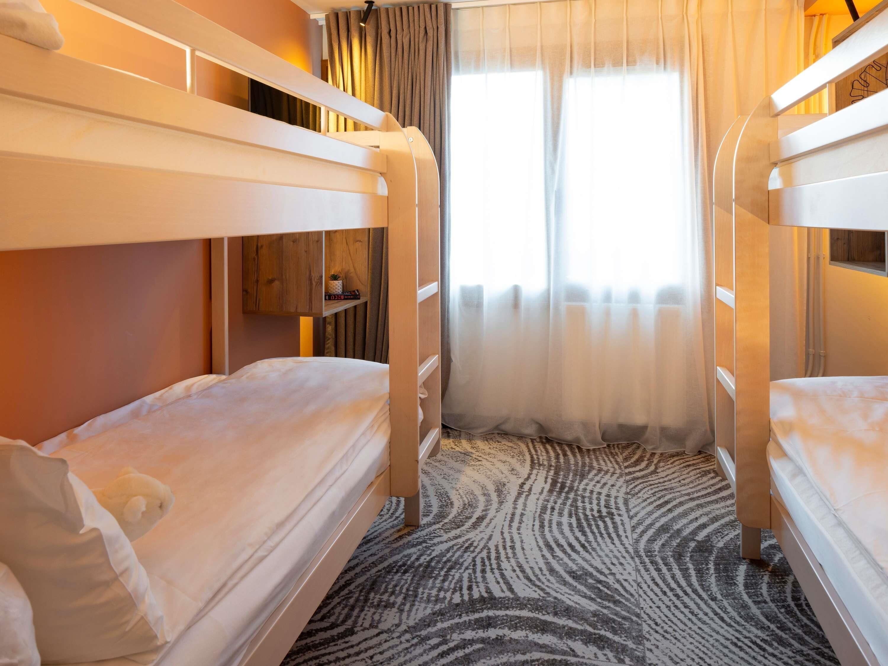A hotel room with bunk beds, a patterned carpet, a wooden shelf with a plant, and a window with sheer curtains.