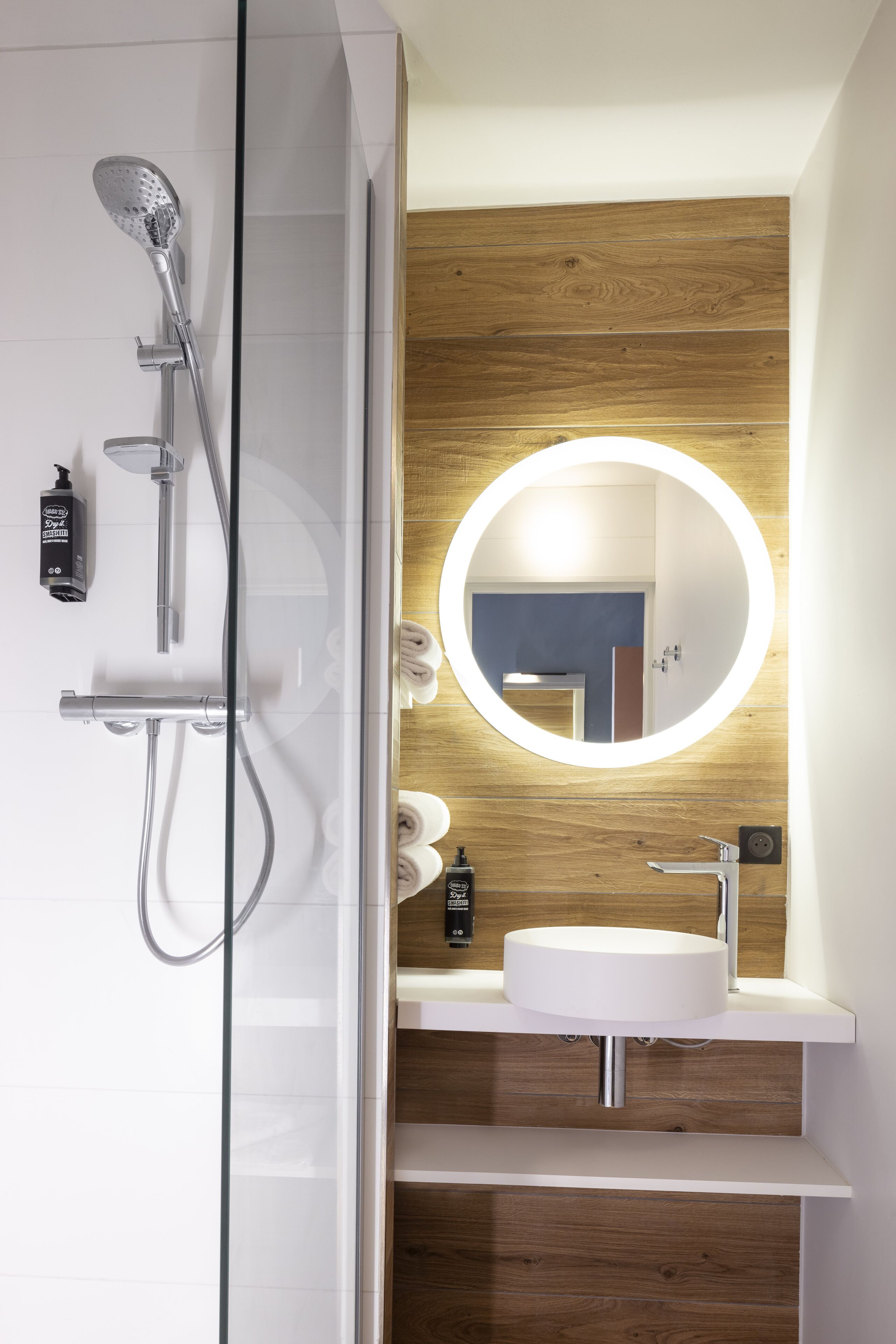 A modern bathroom with a glass shower enclosure, a round mirror with integrated lighting, a white sink, and wooden paneling on the wall.