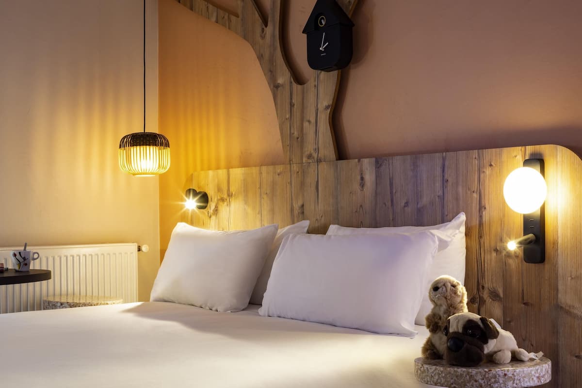 Une chambre à coucher avec une tête de lit en bois, un lit recouvert de draps blancs, une horloge murale et une petite table sur laquelle est posé un ours en peluche.