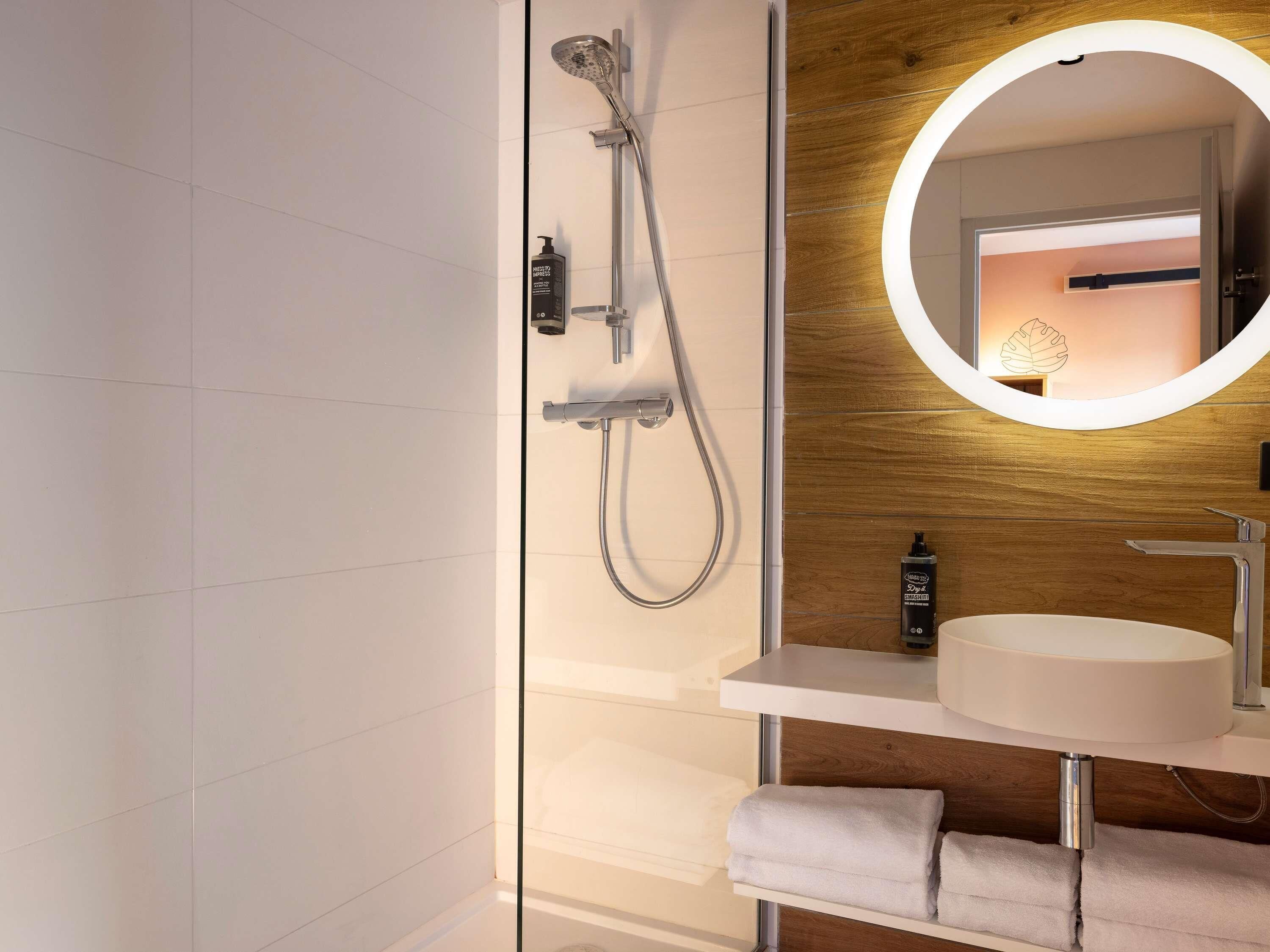 Un baño moderno con cabina de ducha de vidrio, pared de madera, espejo redondo con luz e inodoro blanco.