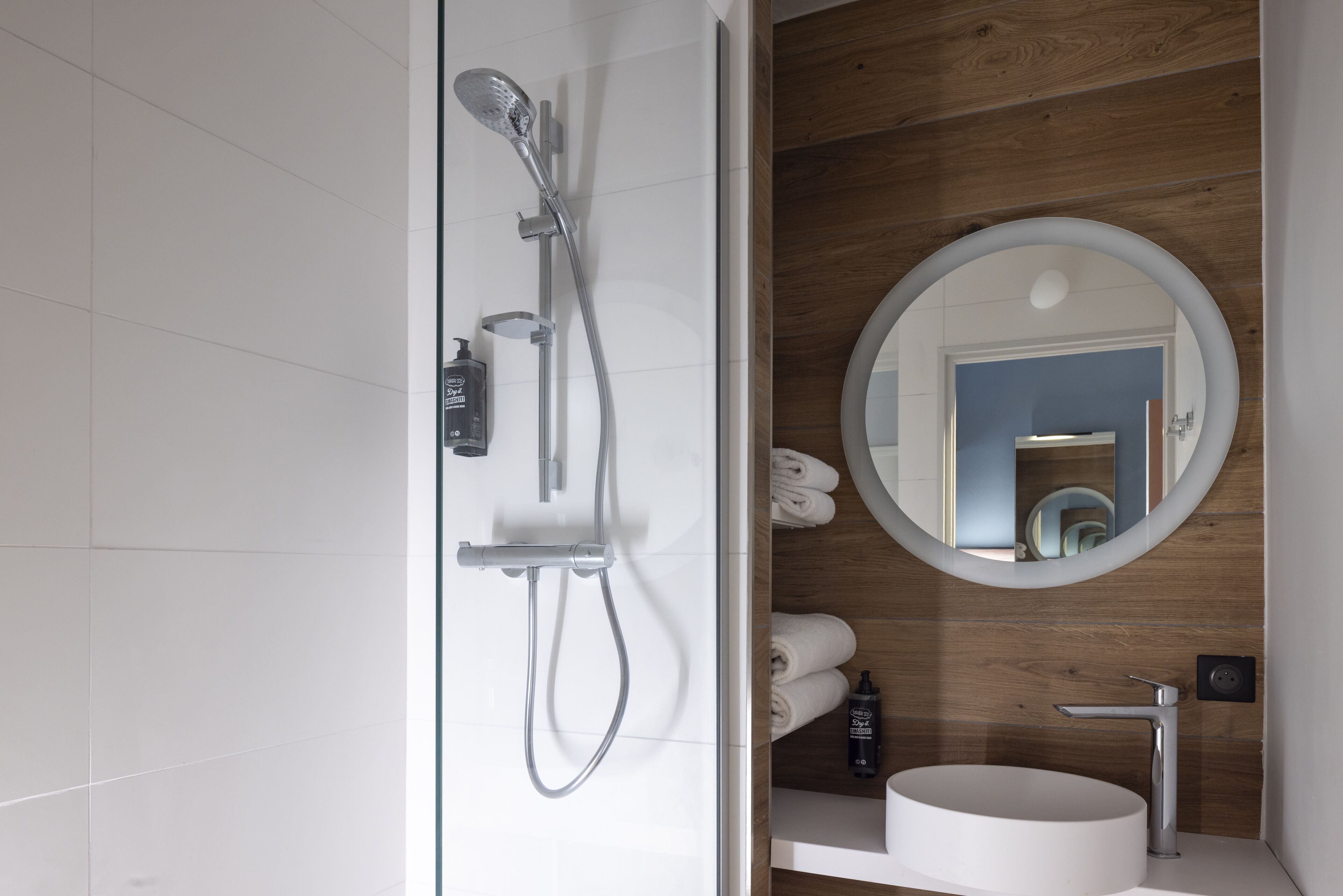 A modern bathroom with a glass-enclosed shower, a round mirror, and a white sink.