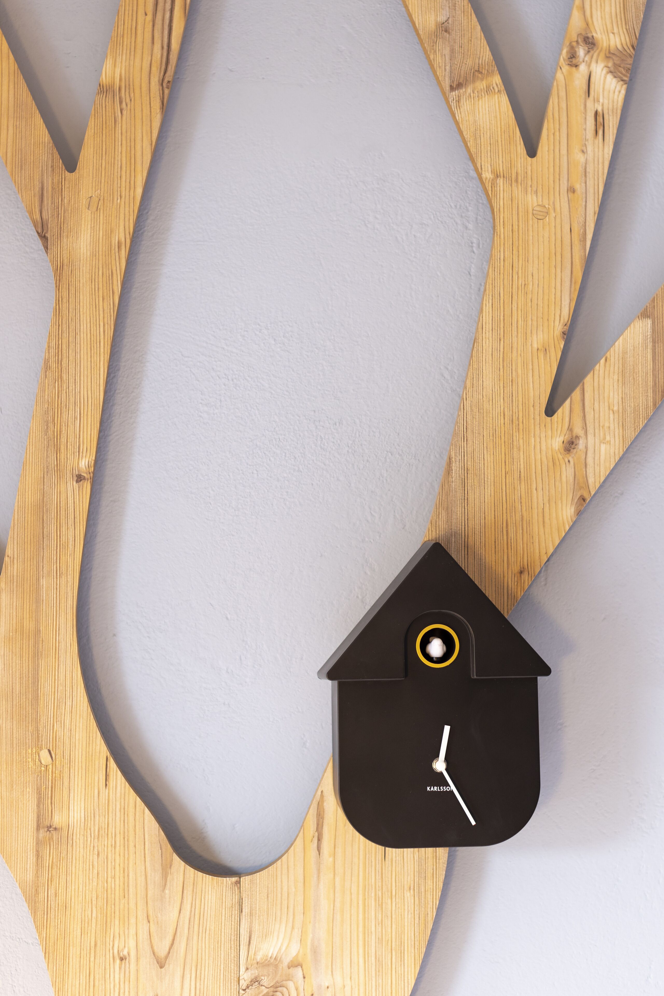 Une pièce moderne et minimaliste, dotée d’un plancher en bois et d’un banc incurvé de couleur grise. Une horloge noire en forme de maison, avec une aiguille des secondes jaune, est fixée au mur.