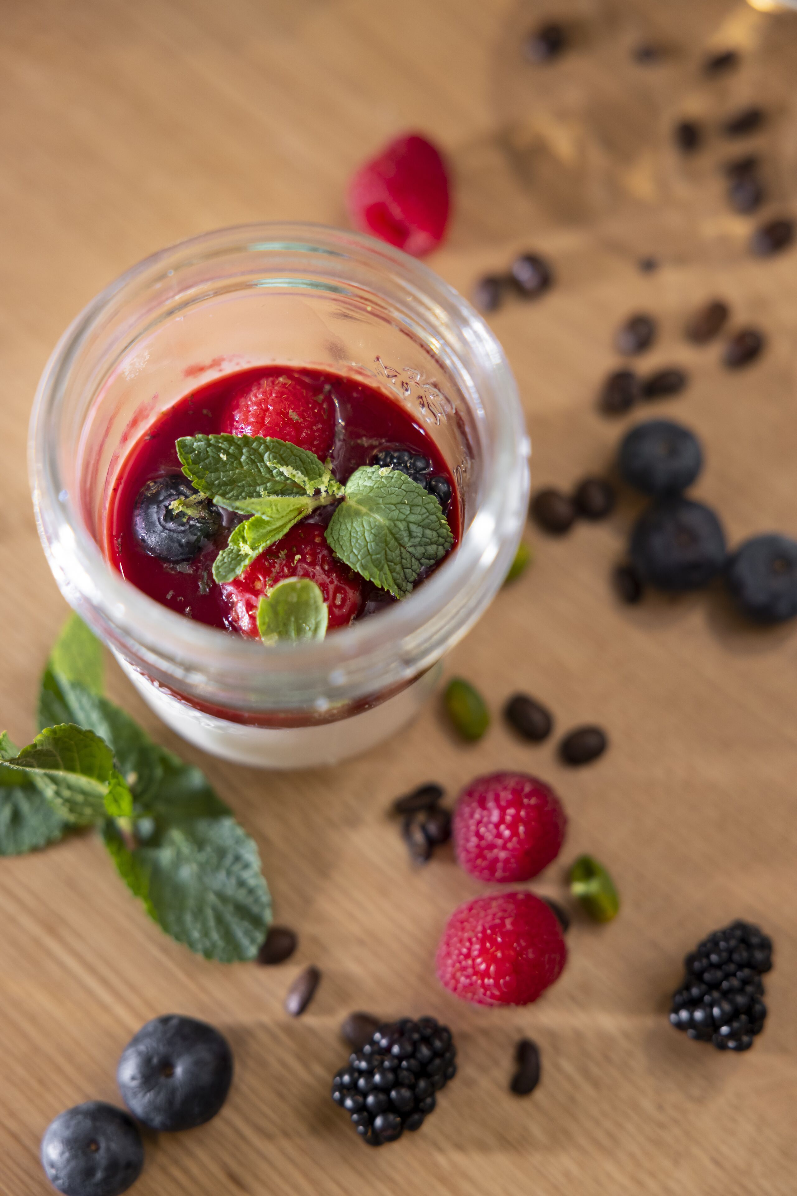 A glass jar filled with a red berry mixture, surrounded by scattered berries and coffee beans on a wooden surface.