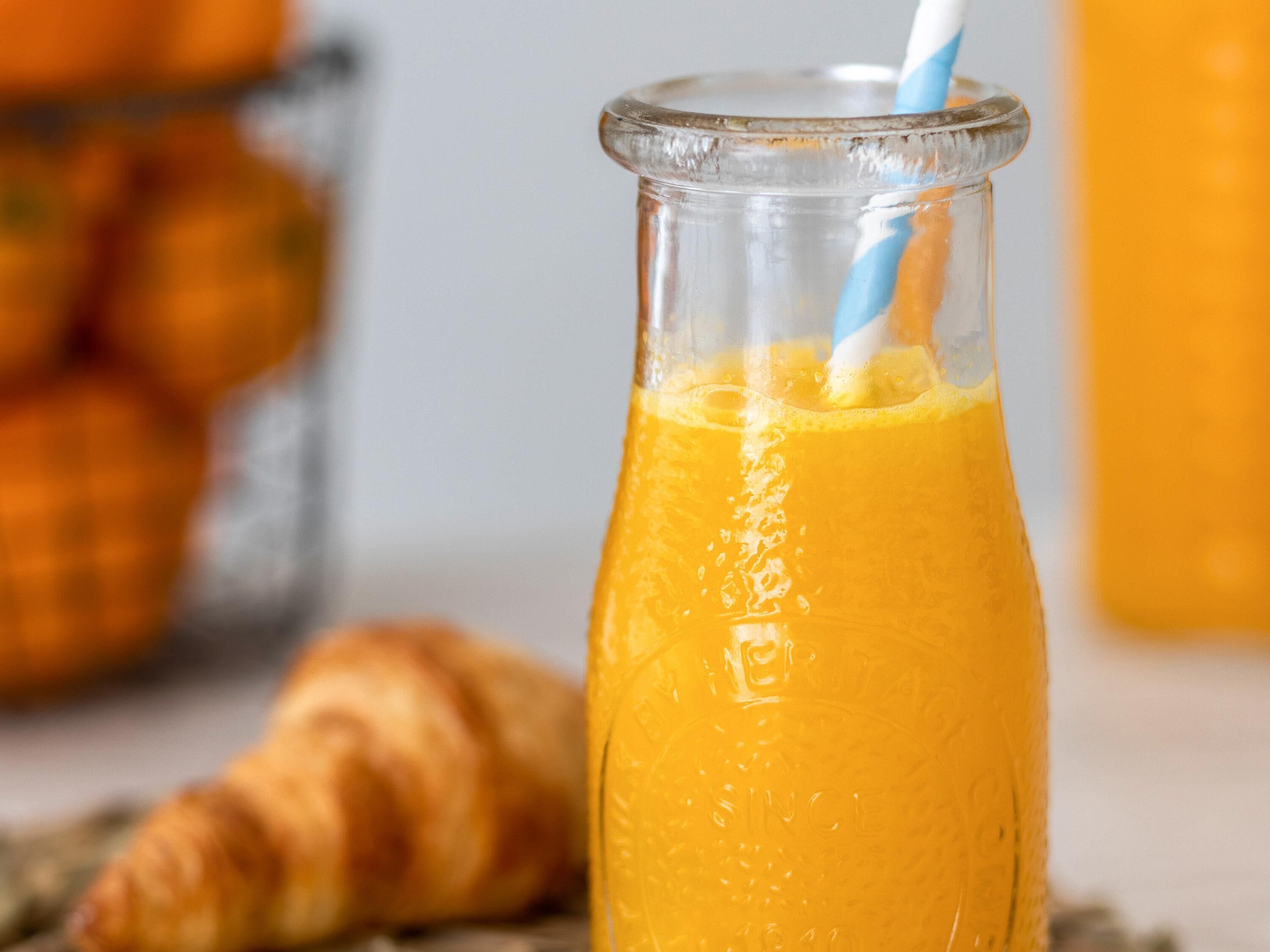 A glass bottle with a blue and white straw filled with orange juice, a croissant, and a blurred background with a glass container.