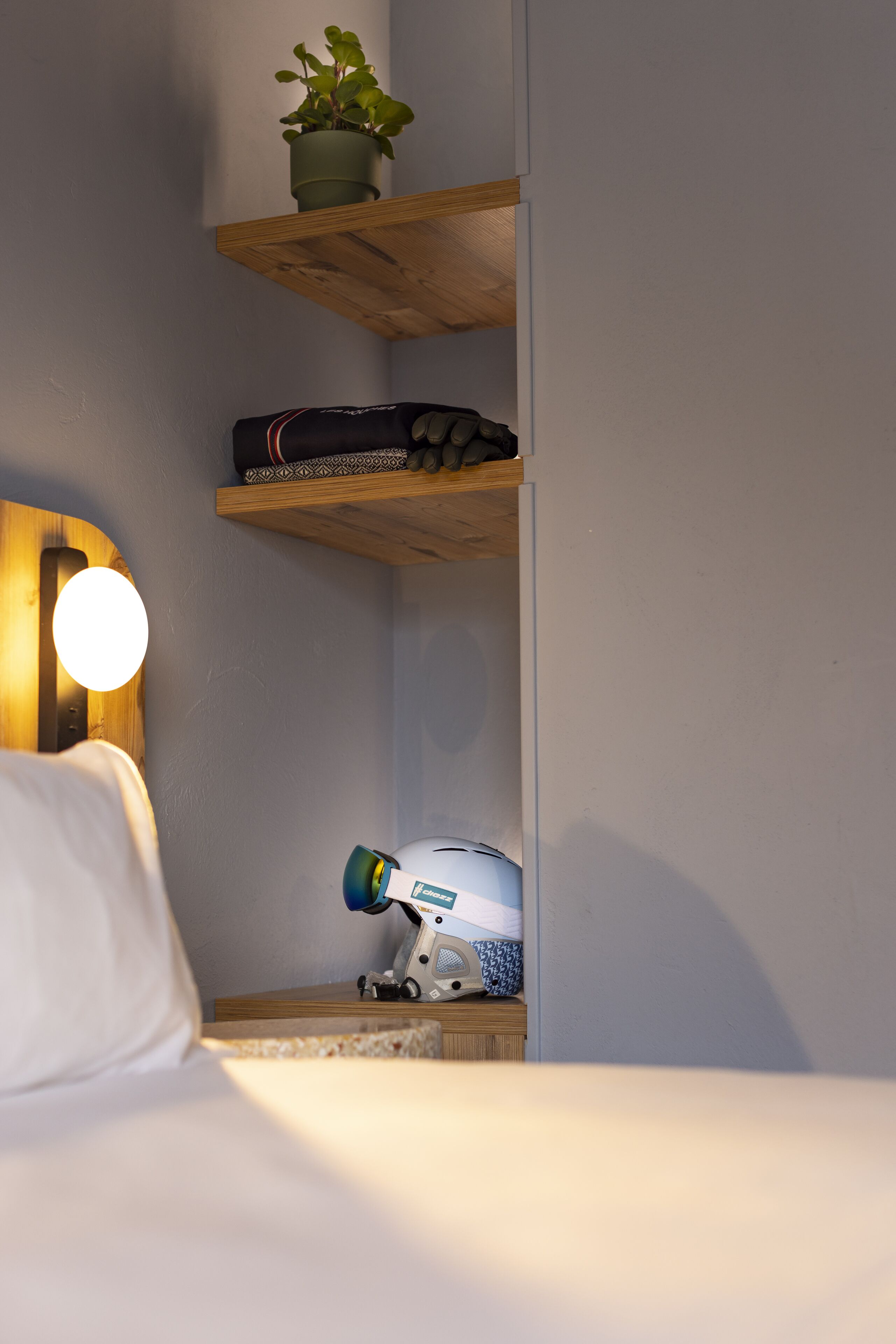 Een hotelkamer met een bed, een bedlamp, een wandplank met een plant, een koffer en een stofzuiger.