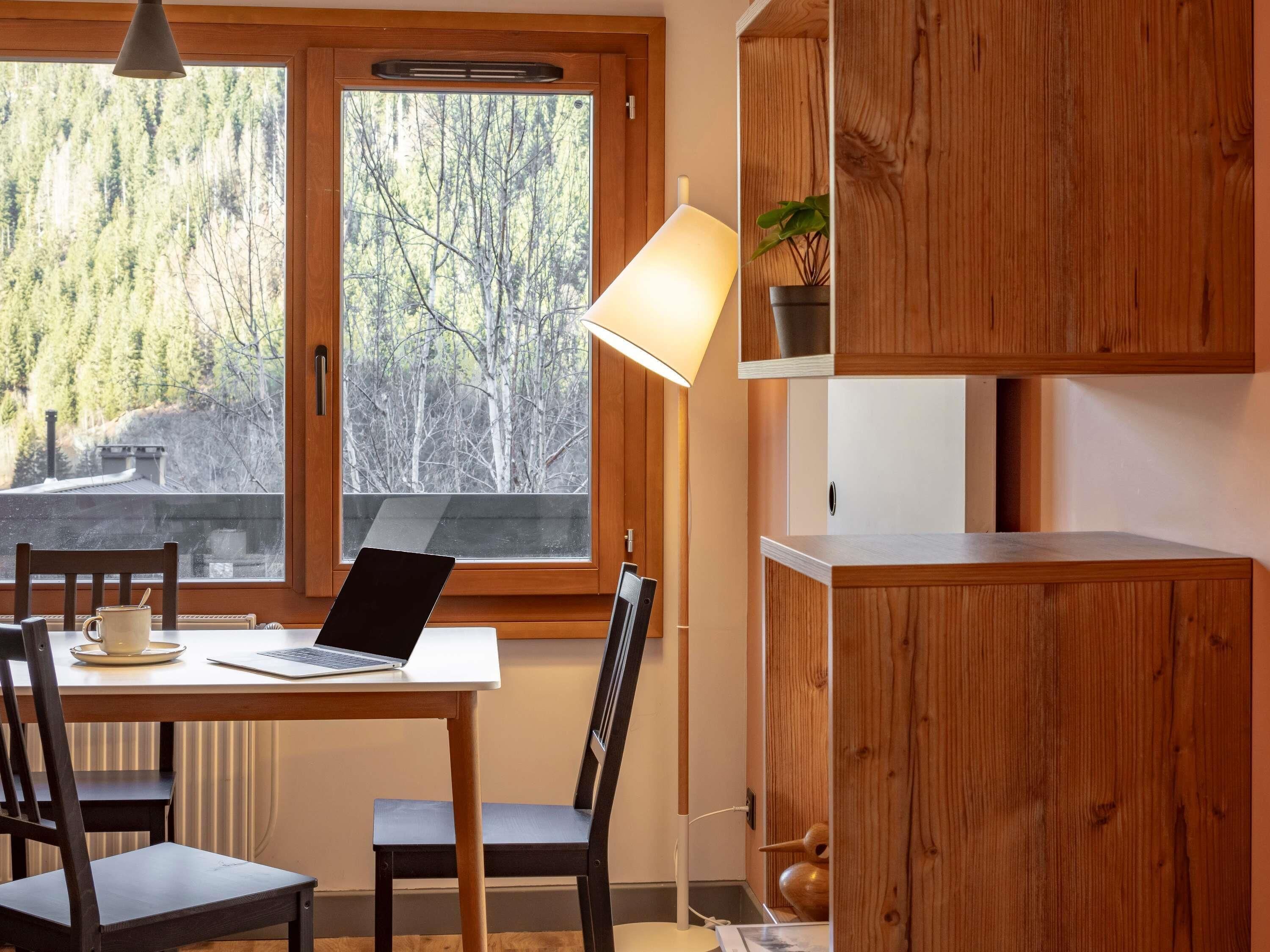 Un interior de madera con una mesa, sillas, una computadora portátil, una lámpara y un ventanal con vistas a árboles.