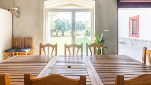 Une table à manger en bois, avec des chaises, un banc et des suspensions vertes décoratives, dans une pièce dont un mur est en béton et qui possède une porte donnant sur l’extérieur.