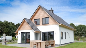Ein modernes, zweistöckiges Haus mit geneigtem Dach, großen Fenstern und einer Terrasse mit Holzmöbeln.