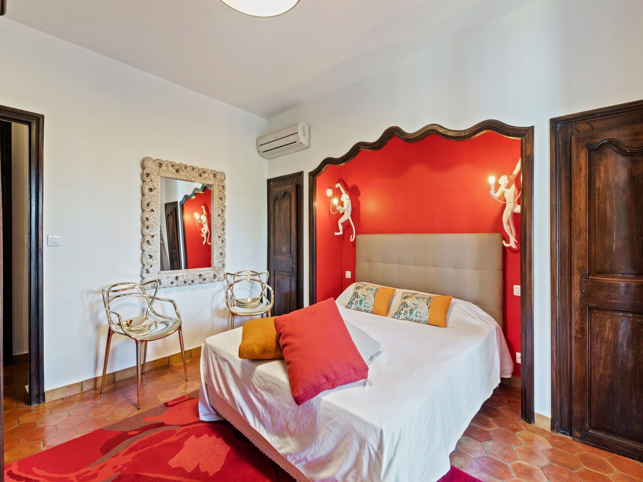 A bedroom with a bed, two chairs, a mirror, and a red accent wall.