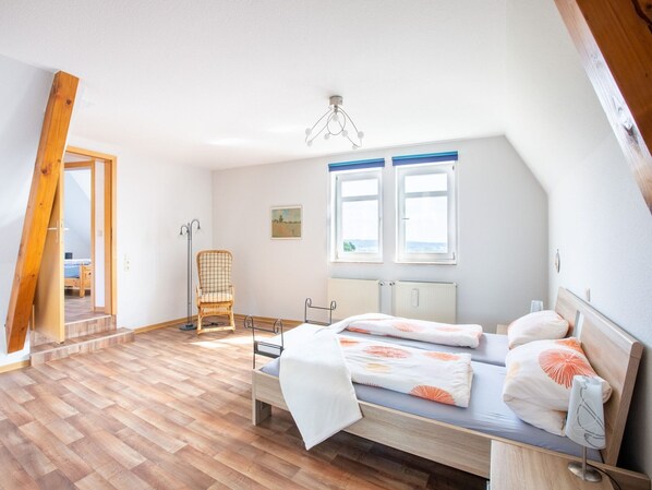 Een slaapkamer met een schuine plafond, houten vloer, een bed met beddengoed met een patroon, een stoel en uitzicht door ramen.
