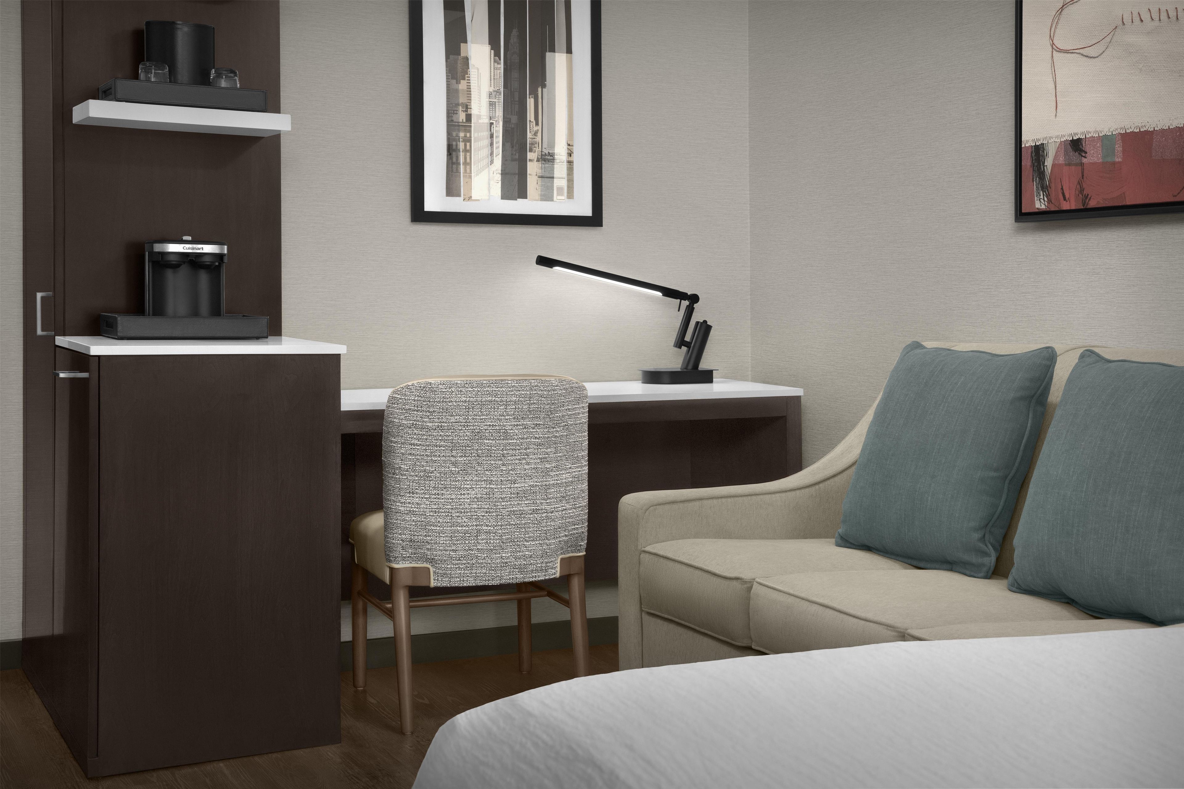 Habitación de hotel con cama, escritorio con cafetera, silla, sofá y una pintura de un paisaje urbano en la pared.